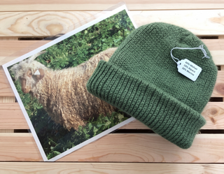 An olive green knit beanie hat with a white product tag, styled on a light wood slatted surface next to a printed photo of a goat.