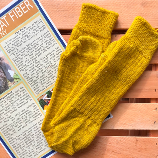 A pair of mustard yellow wool socks laid flat on a wooden slatted surface, partially overlapping a printed informational card about wool.
