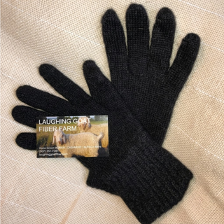 A pair of black knit gloves laid flat on a beige textured fabric background, with a small business card for Laughing Goat Fiber Farm placed over one glove.