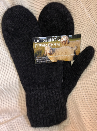 A pair of black knit mittens with a paper tag reading 'LAUGHING GOAT FIBER FARM' placed on a light beige textured fabric background.