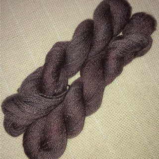 A close-up of two twisted skeins of dark purple yarn resting on a textured beige woven fabric background.