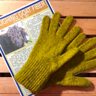 A pair of thick, ribbed knit woolen gloves in a mustard yellow-green color, laid flat on a wooden slatted surface with a magazine underneath.
