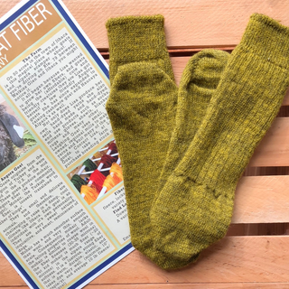 A pair of olive green hand-knit socks laid flat on a light wood slatted surface next to a printed informational card about wool fibers.