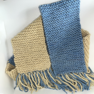 A handknitted scarf with alternating sections of yellow and dusty blue, finished with fringed edges, shown against a plain white background.