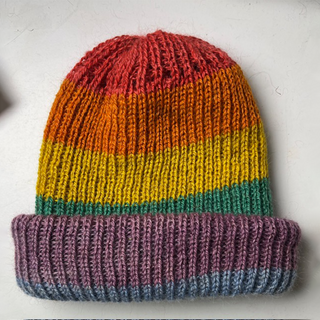 A hand-made knit beanie hat with horizontal rainbow stripes in red, orange, yellow, green, and purple, featuring a folded cuff.
