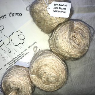 Four balls of natural beige yarn arranged on a gray fabric background, with a paper tag reading '60% Mohair 10% Alpaca 30% Merino' attached to one ball. Part of a branded paper label is visible at the upper left.