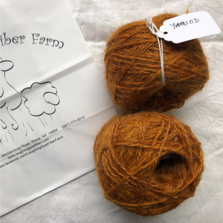 Two balls of coarse, textured yarn in a warm rust-orange color, with one ball tied with a white paper tag. A white envelope printed with 'Angie's Fiber Farm' is visible on the left, set against a white fabric background.
