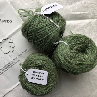 Three balls of sage green yarn with white labels, arranged on a white fabric background next to a paper bag. The labels show the brand 'YMANIS' and a composition of 60% Mohair, 10% Alpaca, and 30% Merino wool.