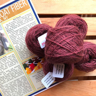Three skeins of burgundy wool yarn resting on a wooden slatted surface next to a printed informational card about goat fiber.