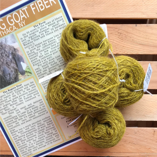Four skeins of thick, textured olive green yarn arranged on a wooden slatted surface next to a printed informational card about goat fiber.