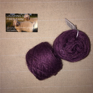 Two balls of deep purple yarn on a beige woven fabric background, with a small 'Laughing Goat Fiber Farm' business card in the upper left and a product tag attached to the right ball.