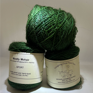Three balls of dark green Mostly Mohair yarn stacked together, with two labels visible. The labels read 'Mostly Mohair' and 'Laughing Goat Fiber Farm'.