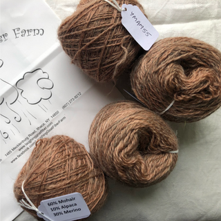 Four balls of brown mohair-alpaca yarn arranged on a light fabric surface next to a printed paper card. The yarn balls have white labels attached, including one that reads YMAM55' and another that reads '60% Mohair 10% Alpaca 30% Merino'.
