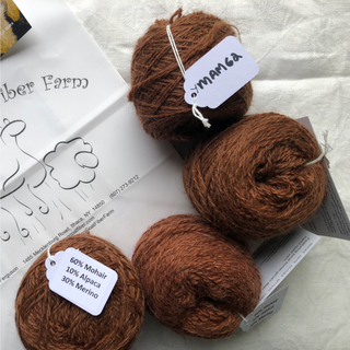 Four brown yarn balls arranged on a white fabric surface with printed paper inserts. Two balls have white paper tags attached; one tag reads 'YMAM62' and another reads '60% Mohair 10% Alpaca 30% Merino.