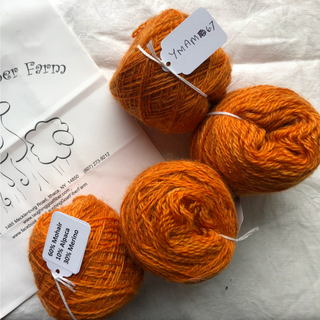 Four skeins of bright orange yarn arranged on a white fabric background next to a white paper card with printed branding and handwritten labels.