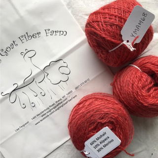 Three balls of coral red mohair alpaca yarn arranged on a white fabric surface next to a white paper bag printed with the Laughing Goat Fiber Farm logo and contact details. Product labels are attached to the yarn balls.