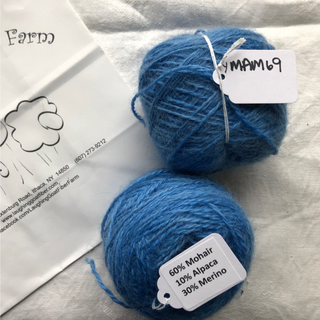 Two balls of blue yarn on a white fabric background, with a paper tag reading '60% Mohair 10% Alpaca 30% Merino' on one ball and a handwritten tag reading 'YMAM69' on the other. A branded paper envelope is partially visible on the left.