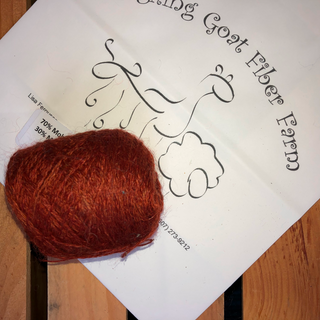 A ball of FLAME-colored wool yarn placed on top of a white paper label that reads 'Laughing Goat Fiber Fiber' on a wooden surface.