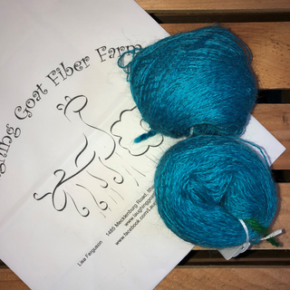 Two balls of bright TURQUOISE blue yarn resting on a white paper card printed with 'Laughing Goat Fiber Farm' and a goat illustration, placed on a wooden slatted surface.