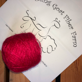 A bright red ball of yarn placed on top of a white printed card with black script text reading 'Laughing Goat Fiber Farm' on a wooden surface.